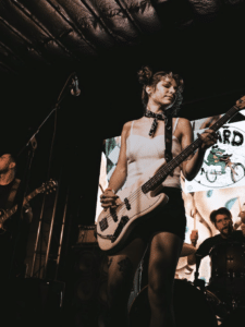 A woman plays an electric bass guitar on stage with a band at festivals, while a drummer and another guitarist perform in the background.